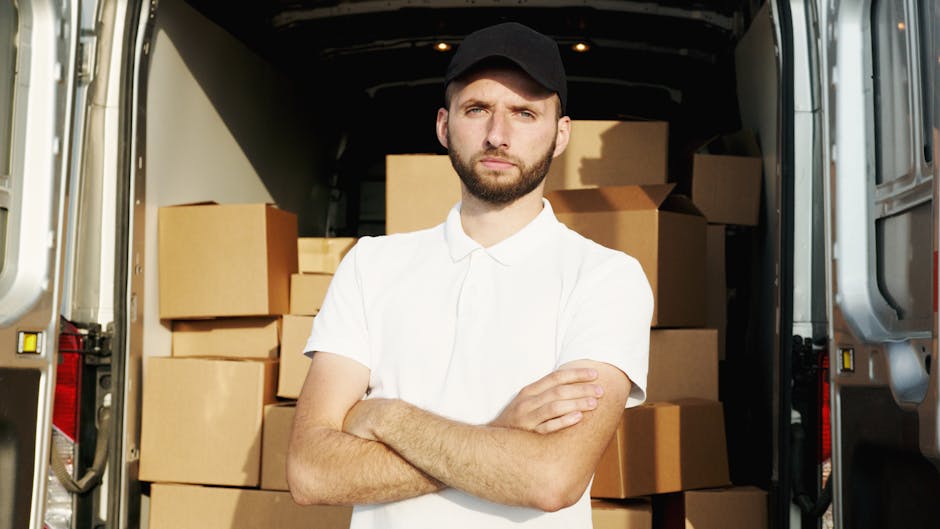 A man standing inside the back of a moving van, surrounded by numerous packed cardboard boxes of various sizes. The boxes are stacked neatly against the van's interior walls, some secured with plastic