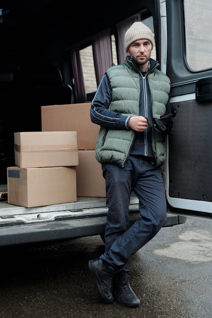 A man standing inside the back of a moving van, surrounded by numerous packed cardboard boxes of various sizes. The boxes are stacked neatly against the van's interior walls, some secured with plastic