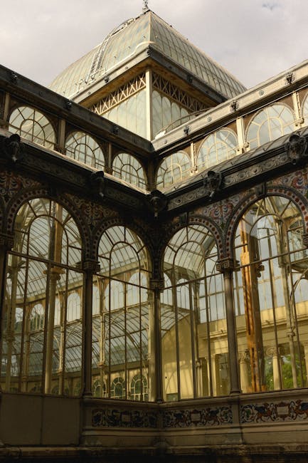 Close-up view of an ornate Victorian-style glass greenhouse with a decorative metal framework painted black, featuring large arched windows and intricate detailing. The structure includes a prominent corner section with a pyramidal glass roof, revealing glimpses of the interior. The greenhouse is situated outdoors under an overcast sky, with natural light reflecting off the glass panels. This architectural detail may be part of a historic property undergoing house removals or furnishing transport, as seen in professional relocation services like those offered by Man with Van Crystal Palace. The image emphasizes the craftsmanship of the glass and metal construction, which is typical of period structures involved in home relocation and furniture transport projects.