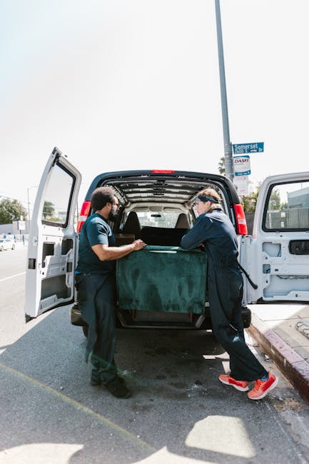 Two movers from Man with Van Crystal Palace are engaged in a home relocation process, loading a large, green upholstered furniture piece into the back of a white cargo van parked on a paved urban street. Inside the van, visible through the open rear doors, are empty space and some protective padding. Both movers are wearing dark uniforms and are carefully lifting the furniture, which is positioned on a dolly or hand truck for easier handling. The scene is set outdoors during daylight, with a street sign visible on a pole and a clear sky overhead. Nearby, a curb with a pinkish concrete edging indicates the loading area, and the presence of other parked vehicles suggests a typical city environment. This image reflects a furniture transport and loading process typical of professional house removals, supported by the services of Man with Van Crystal Palace in the Westow Hill area.